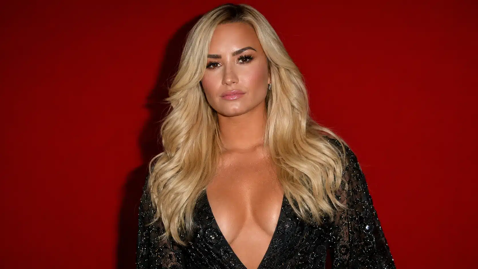portrait of demi lovato after overdose journey showing blonde hair in a black shimmery dress against a red backdrop