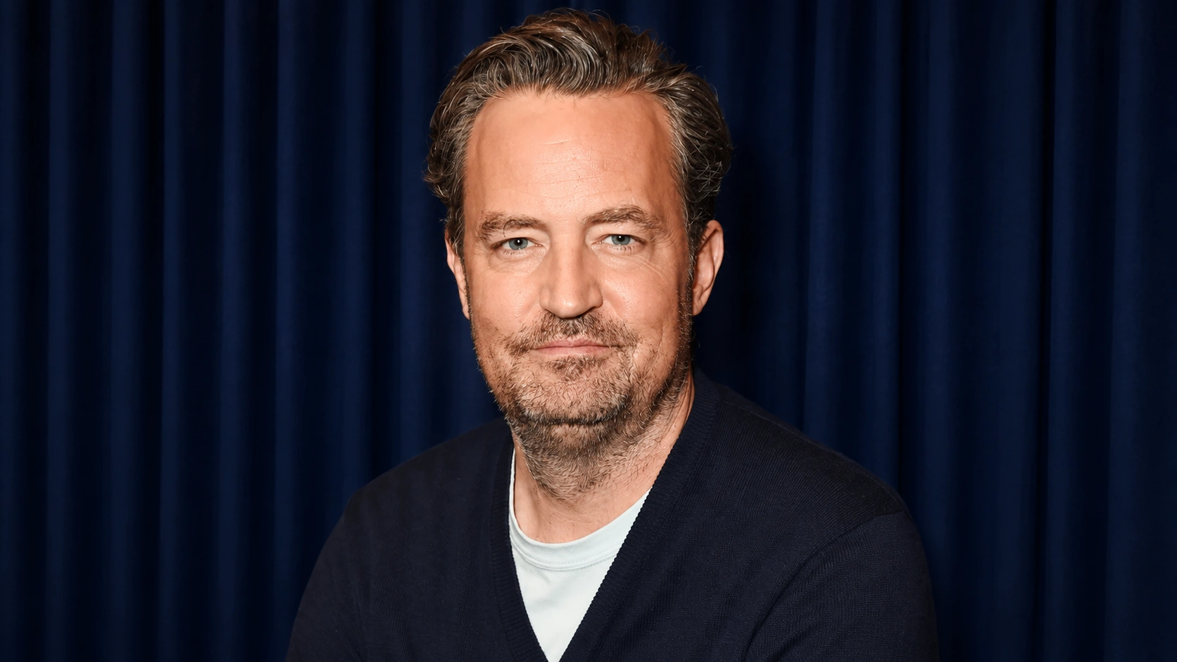 portrait of matthew perry with short hair and light beard wearing a dark sweater against a dark blue curtain background