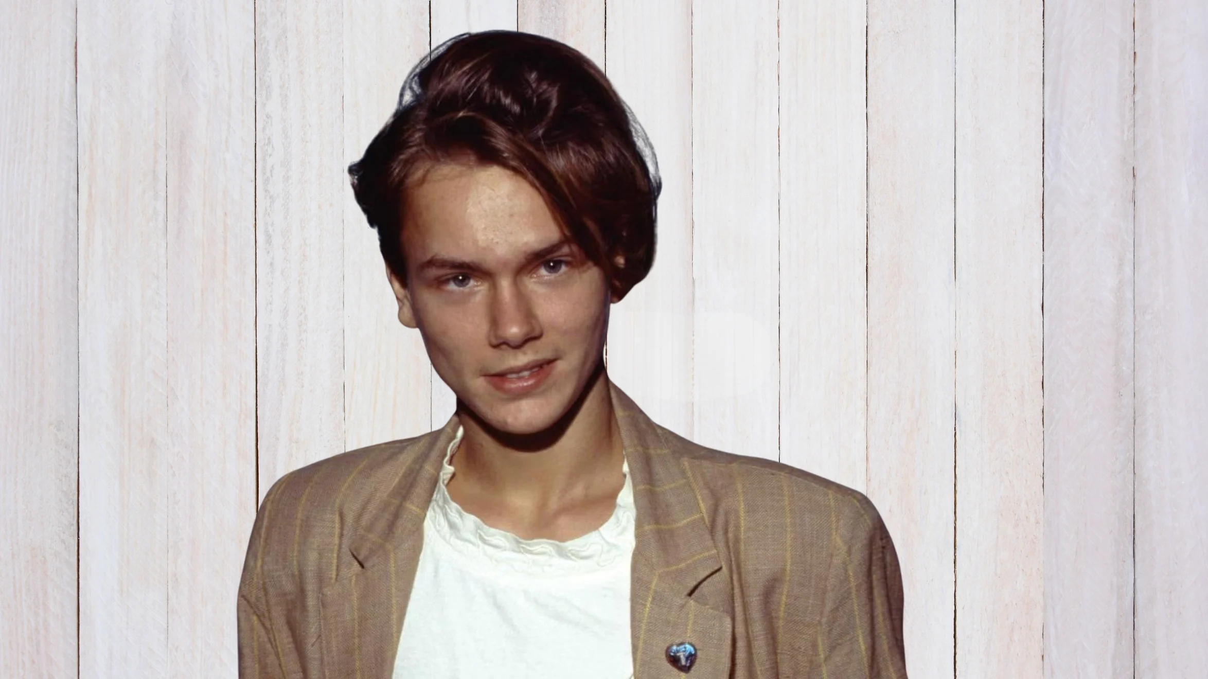 portrait of person with short styled hair wearing blazer and white shirt, standing against light wooden panel background