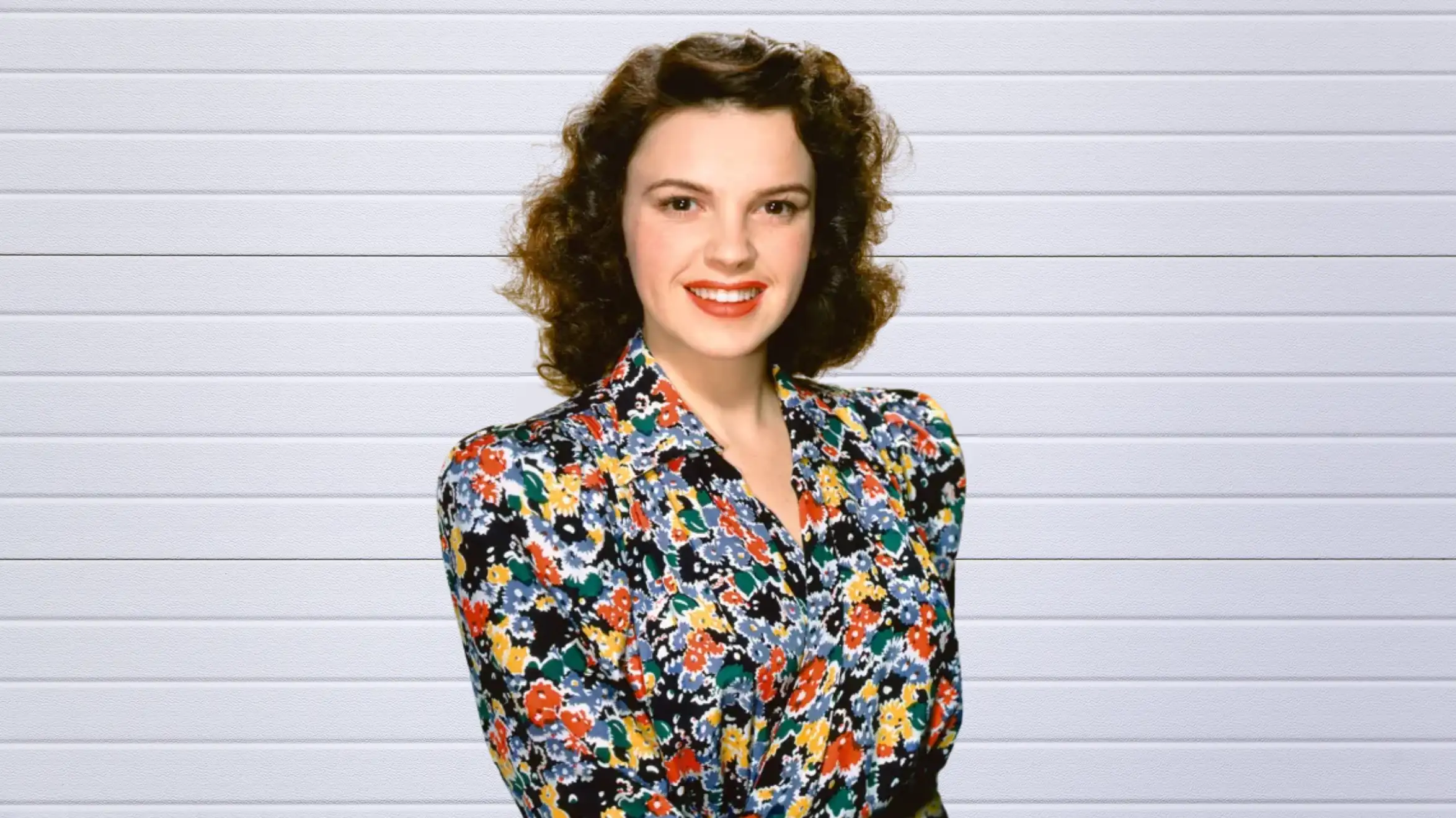 portrait of smiling woman with curly hair wearing colorful floral blouse, standing against white paneled background
