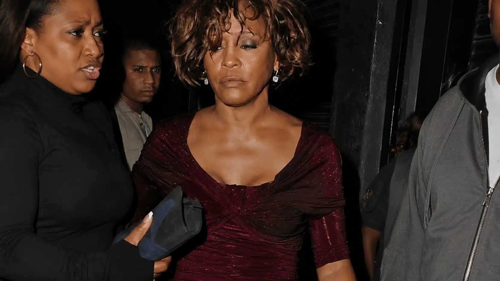whitney houston in a dark red shimmering dress, with short curly hair, escorted by two people at a night event