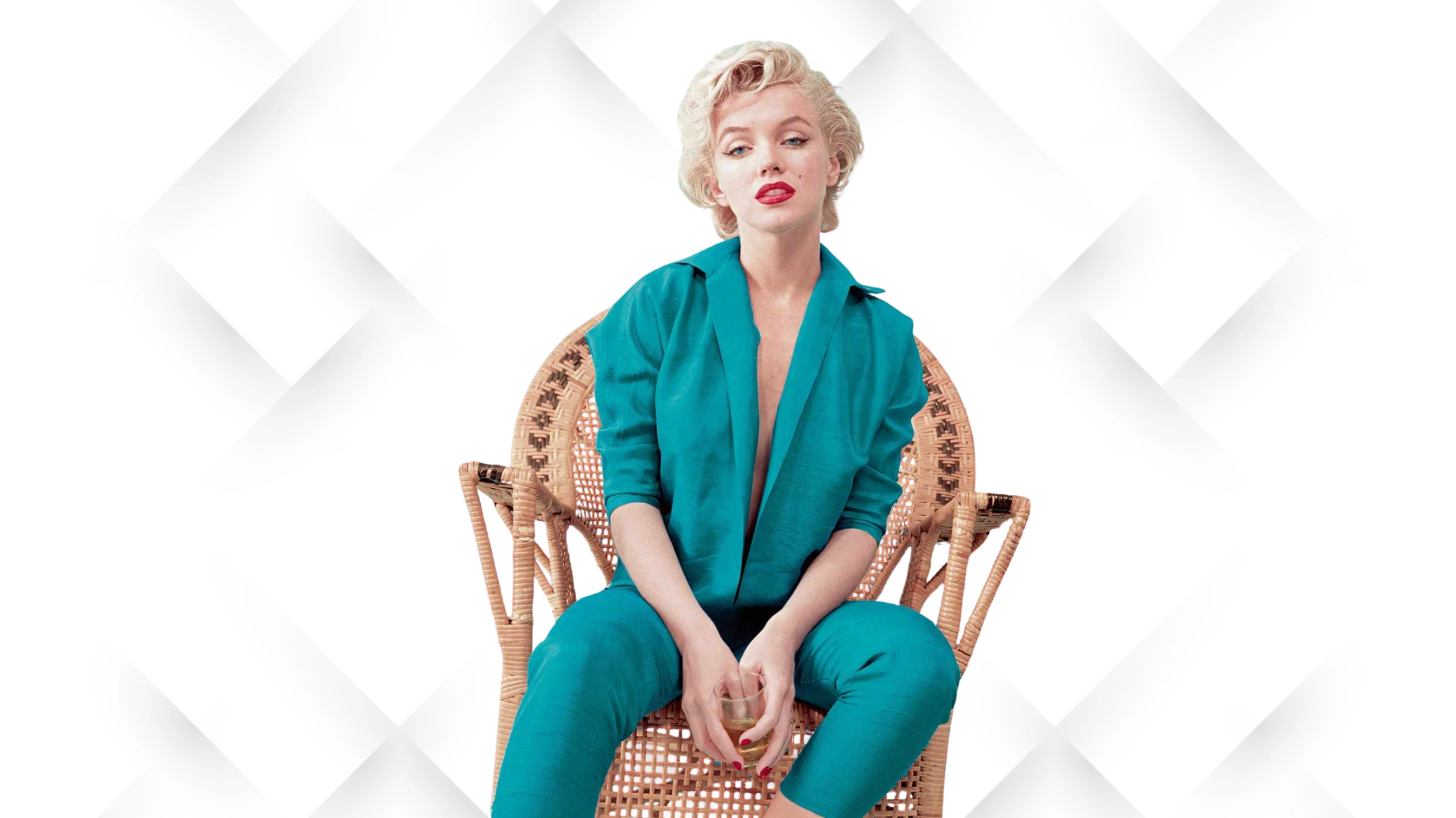 woman in teal outfit sitting on wicker chair holding glass, against light patterned background, looking at camera