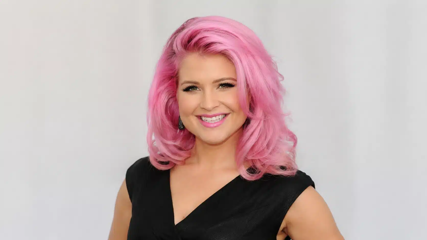 woman with voluminous pink hair, wearing black dress, posing against a smooth gray background, confident and stylish