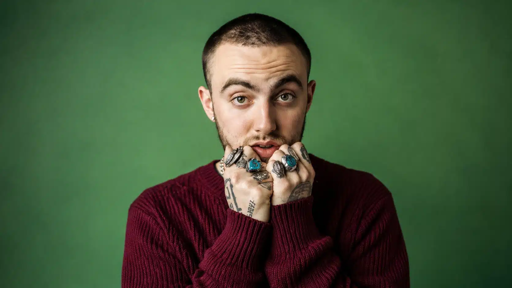 young man in a maroon sweater with tattoos and rings, holding his hands near his mouth, against a green background
