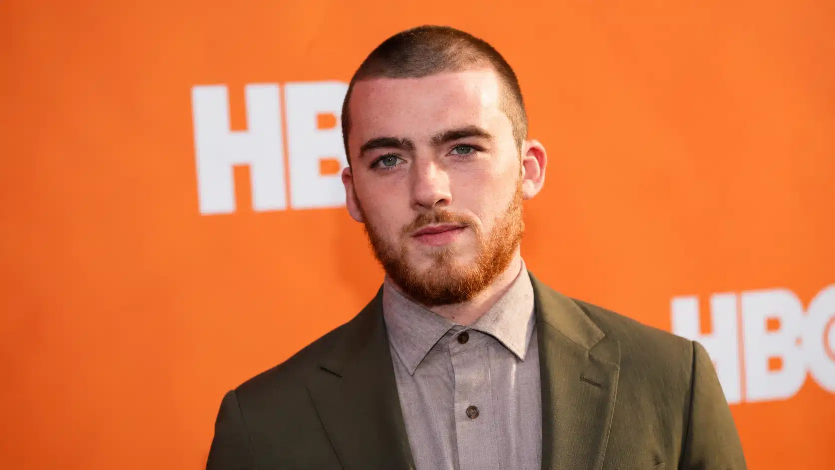 young man with a buzz cut and beard, wearing an olive green blazer and gray shirt, standing in front of an orange banner