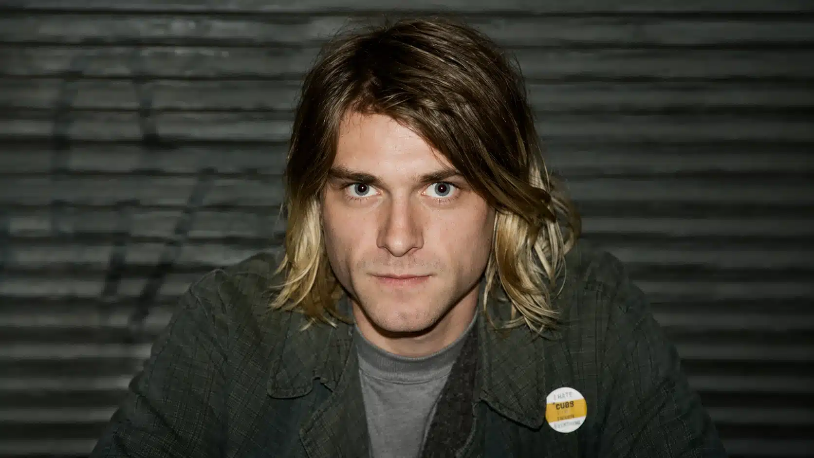 young man with blonde hair, wearing a gray shirt and plaid jacket, sitting in front of a metal shutter backdrop