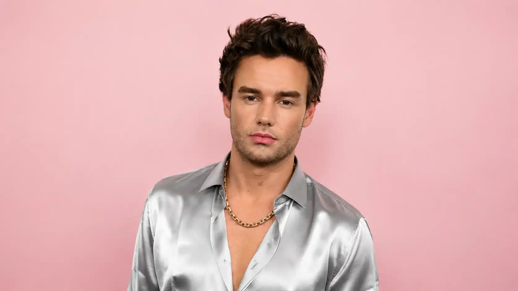 young man with tousled hair wearing a silver shirt and chain necklace, standing in front of a baby pink background