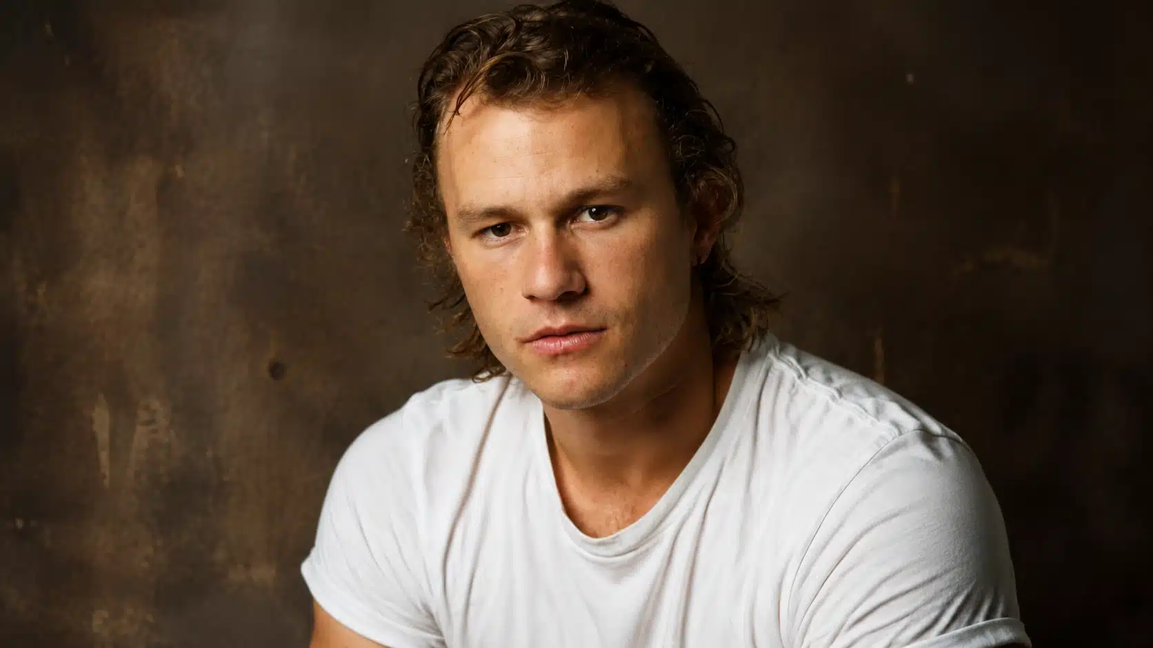 young man with tousled hair, wearing a white t-shirt, gazing intensely at the camera against a dark brown background