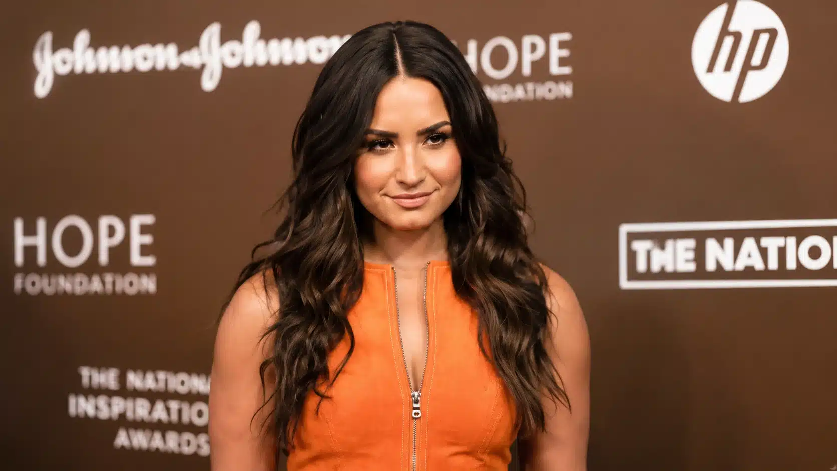young woman with long wavy hair wears orange dress, posing confidently against brown backdrop with white logos at event