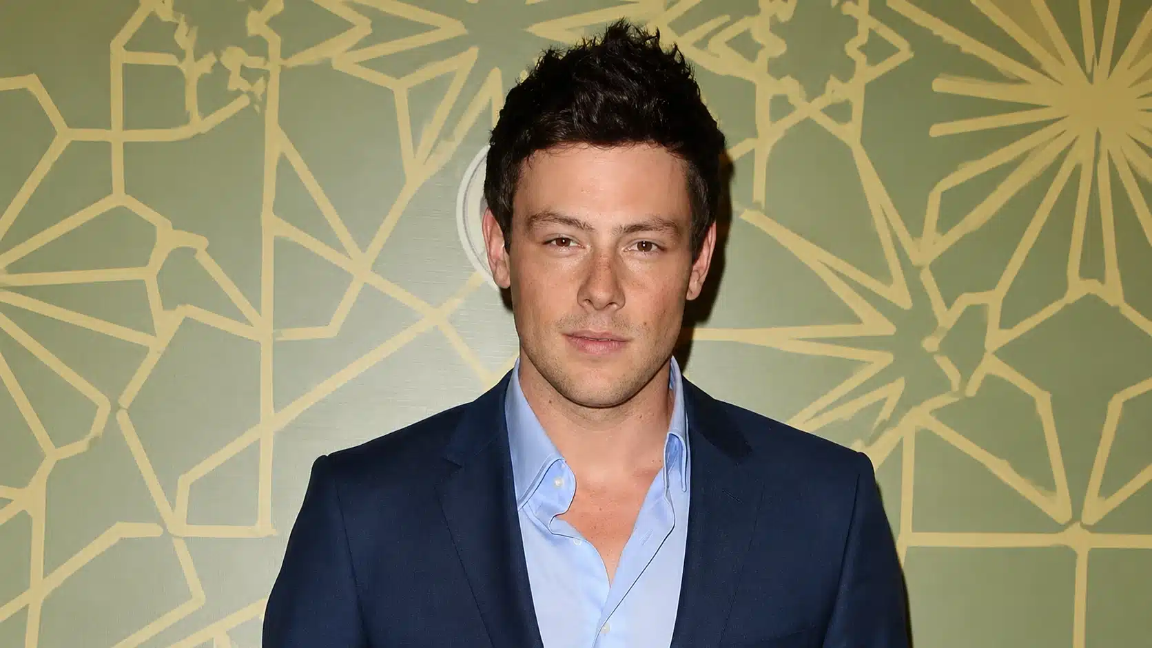 a man in a navy blazer and light blue shirt stands before a green geometric patterned backdrop, facing the camera