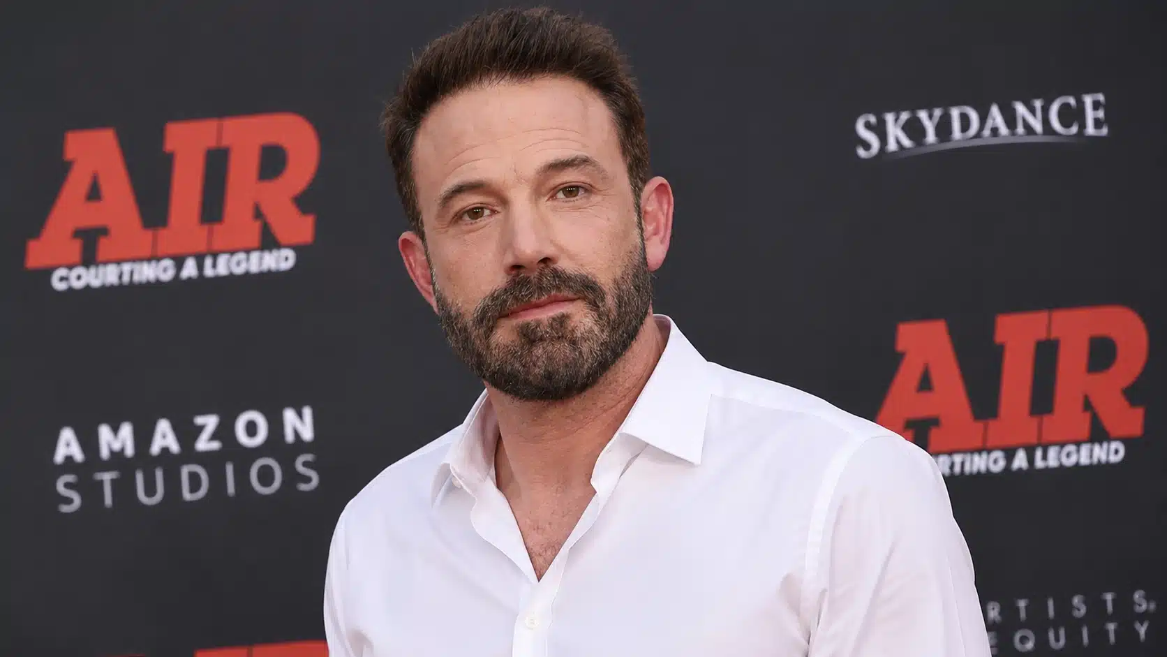 bearded man in a white open-collar shirt poses at a red-carpet event before a dark branded movie backdrop