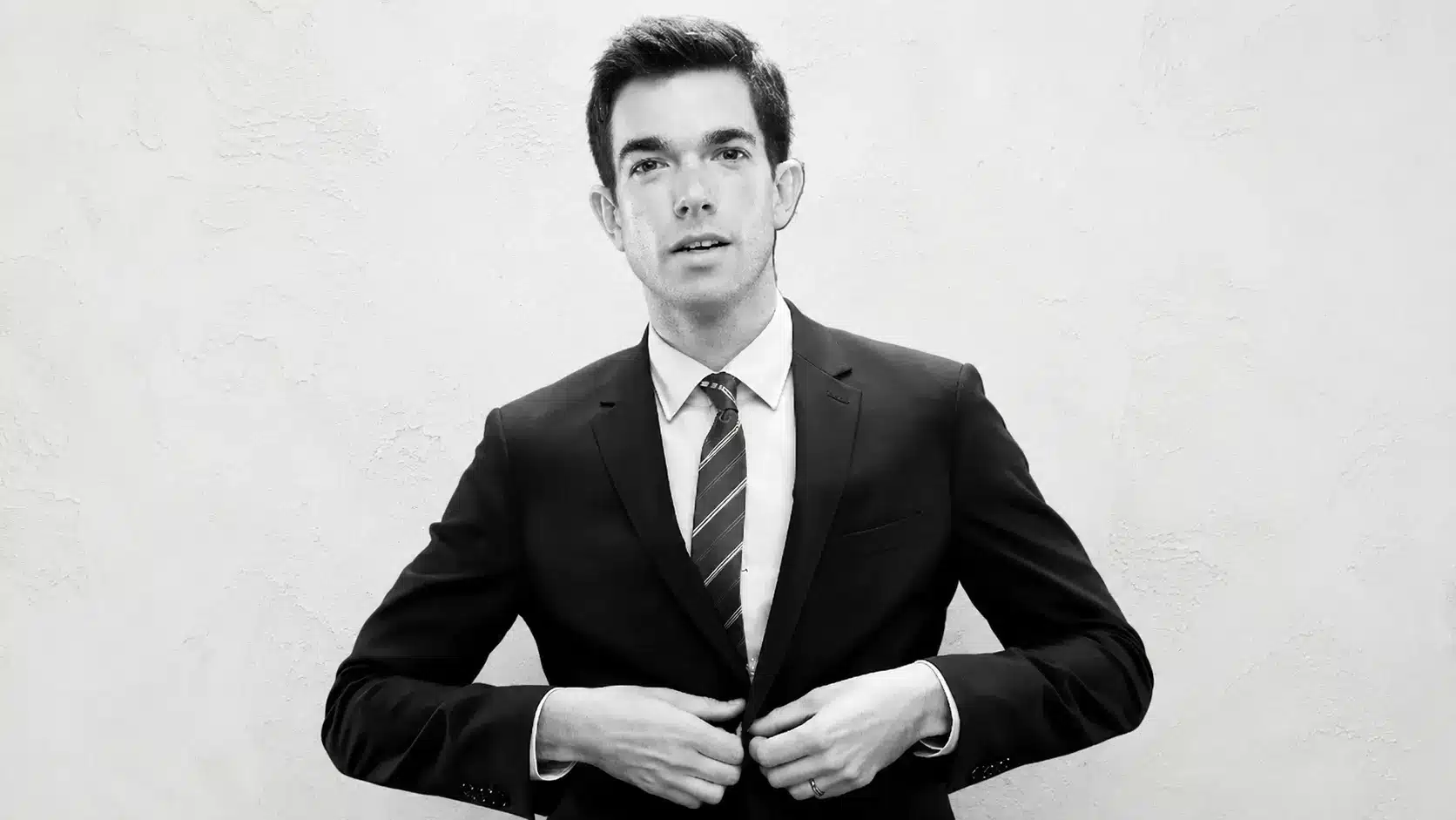 black and white portrait of john mulaney in a suit standing against a rustic white textured background.