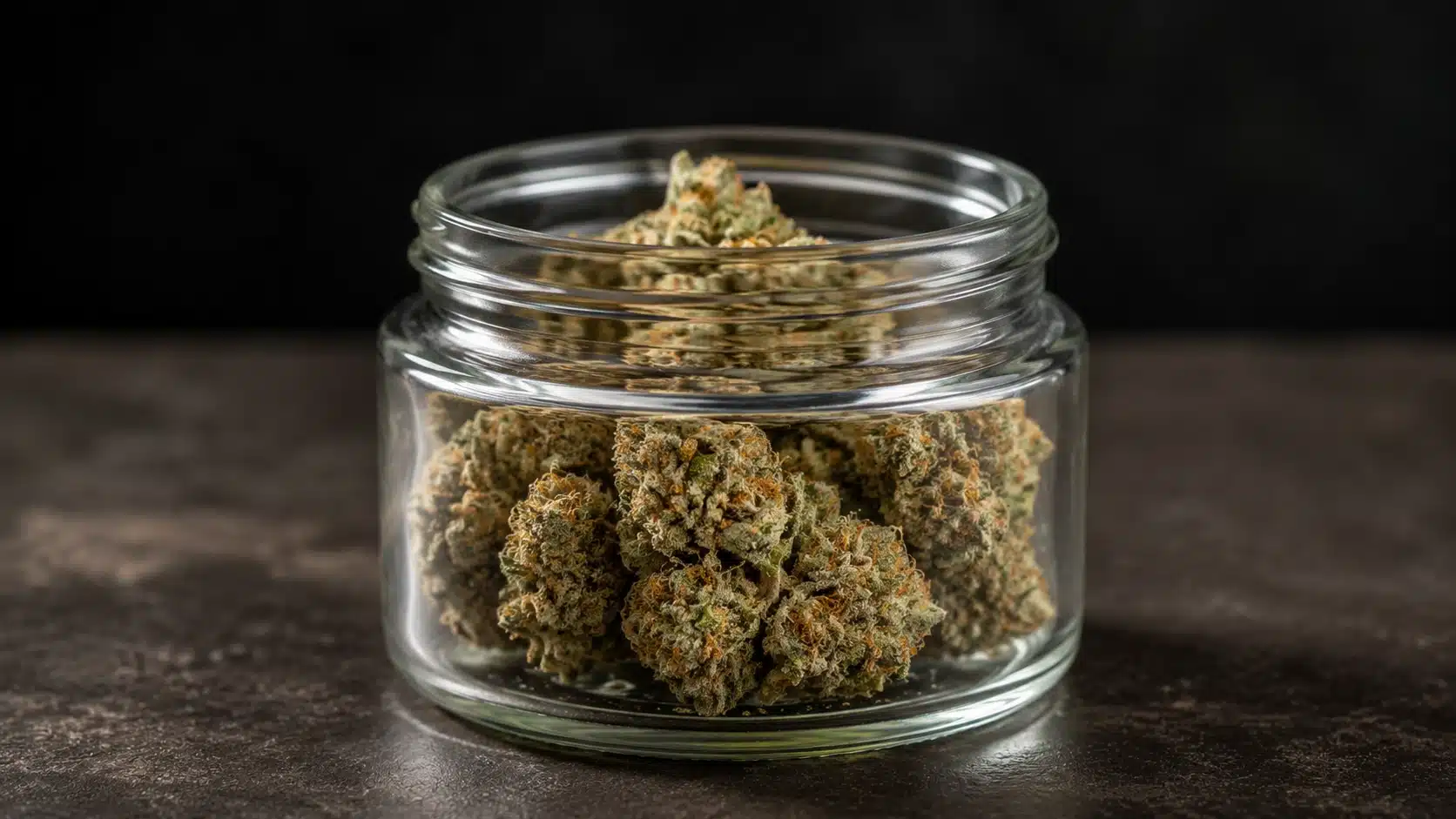 clear glass jar filled with frosty gg4 cannabis buds on a dark table with a plain black wall behind it