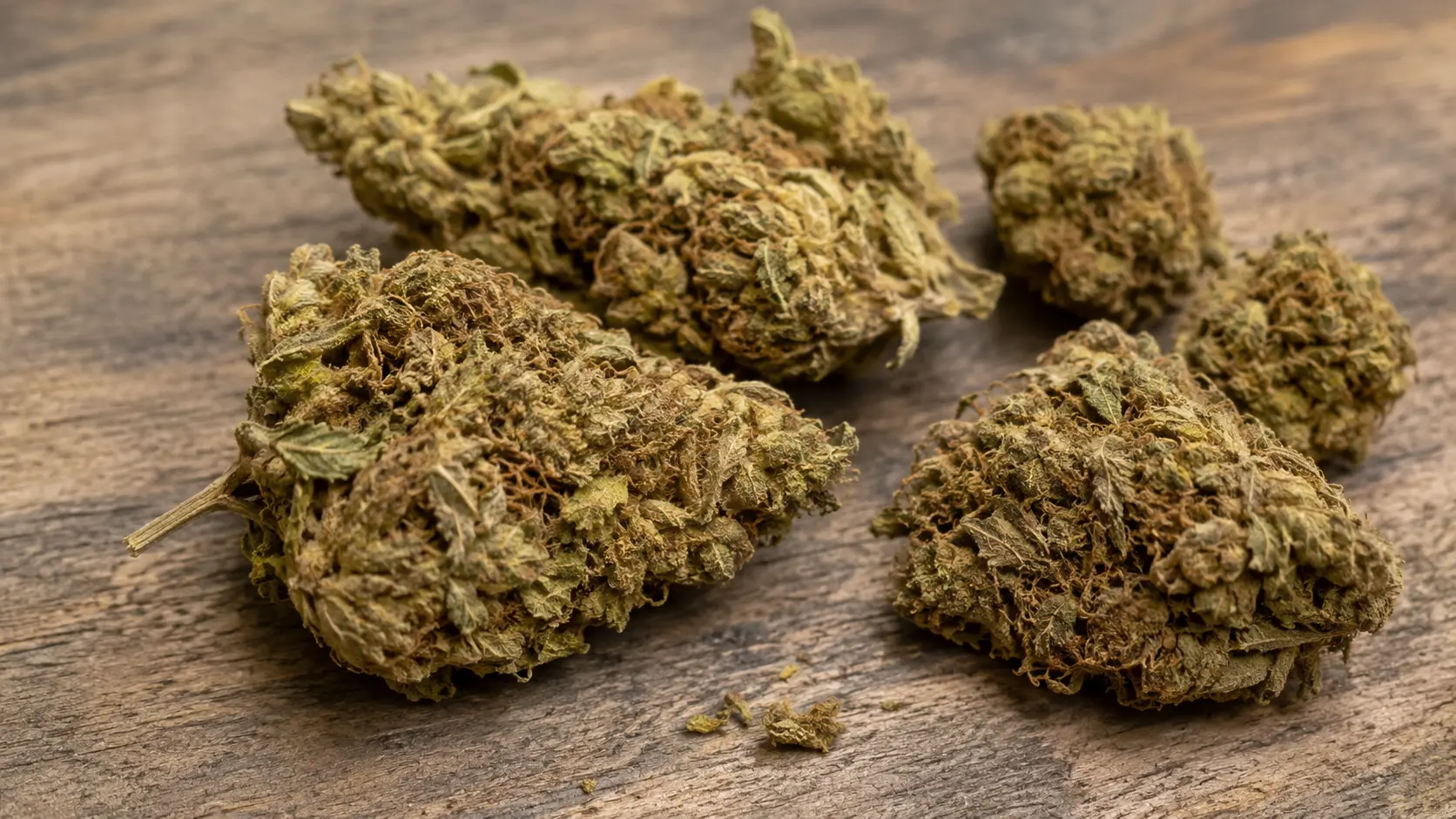 close-up of dry cannabis buds on a rustic wooden surface, showing brown-green texture, stems, and curled leaves