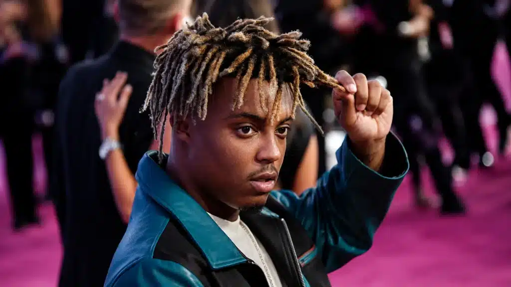 close-up portrait of a man in teal jacket holding blond-tipped hair on a pink carpet with blurred event crowd behind