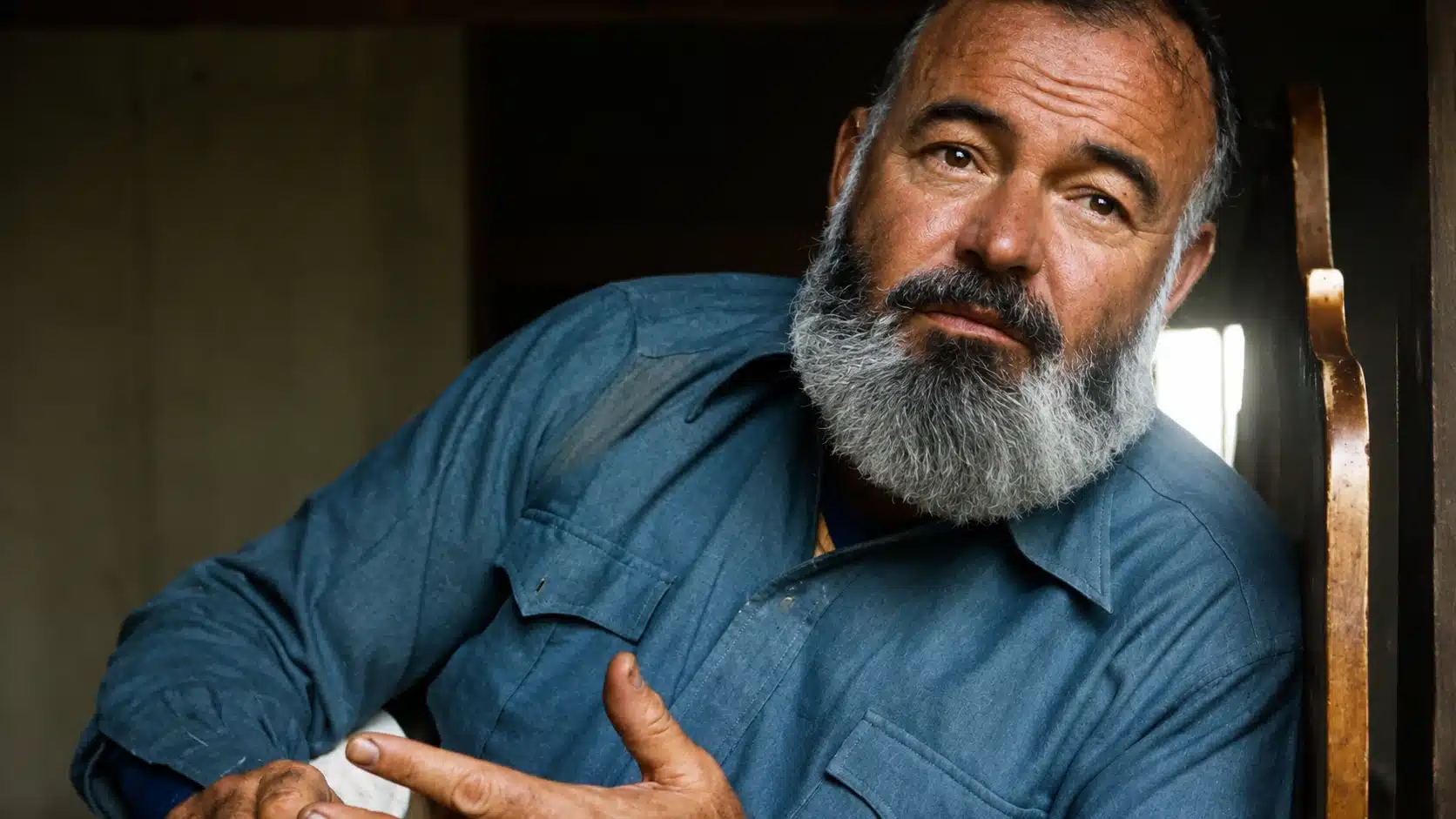 colorized close up portrait of a bearded man in blue shirt leaning indoors beside a bright window with warm light
