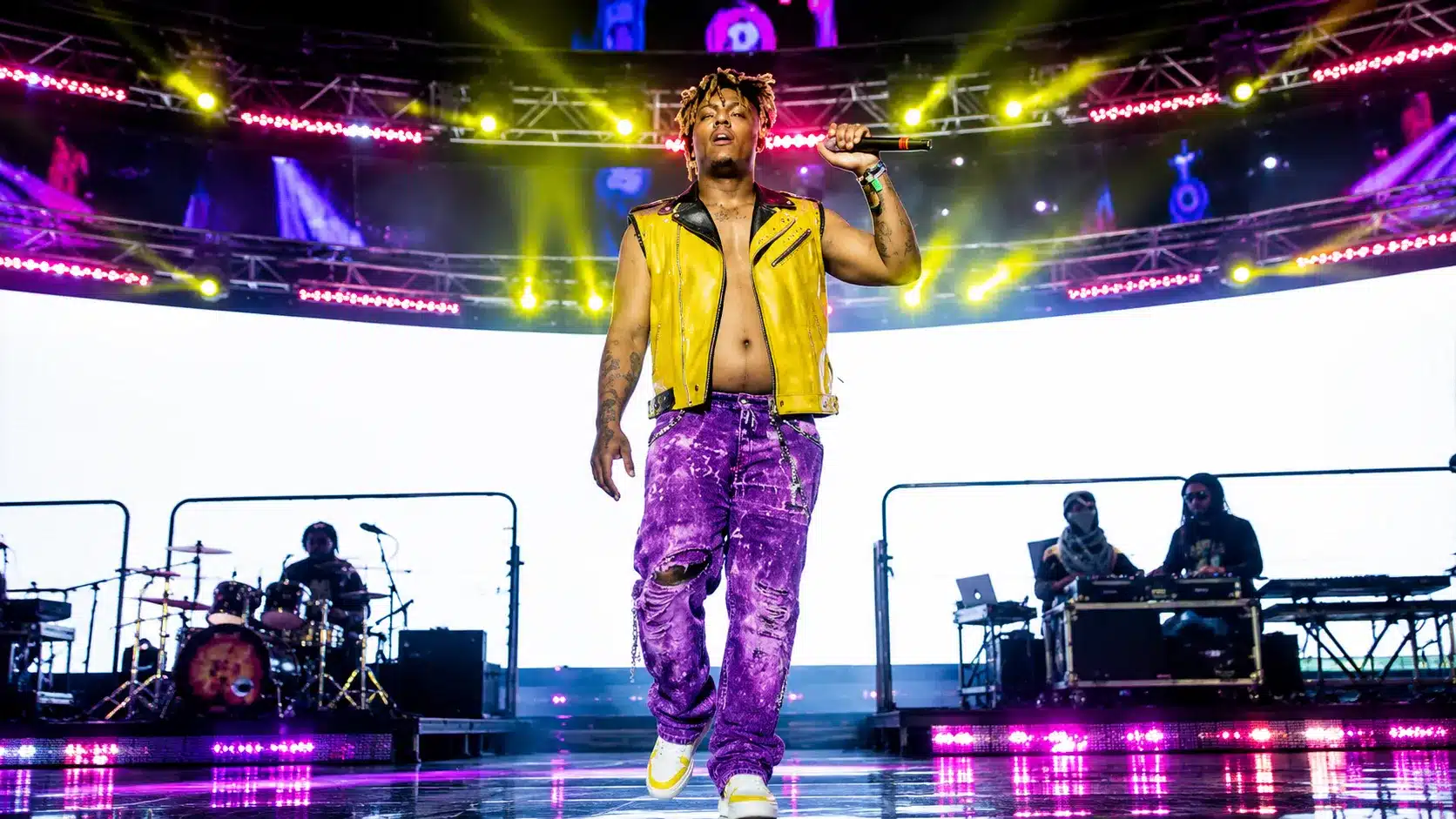 concert performer in yellow vest and purple pants holding a microphone on a bright stage with band members behind