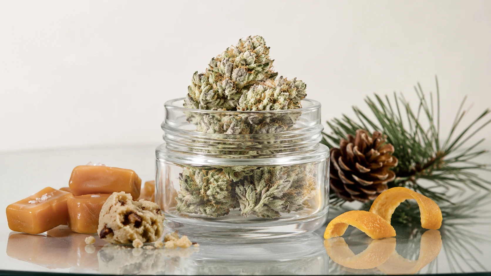 glass jar of frosty cannabis buds on glass table with caramel, cookie crumbs, pinecone, needles, and orange peel flavors