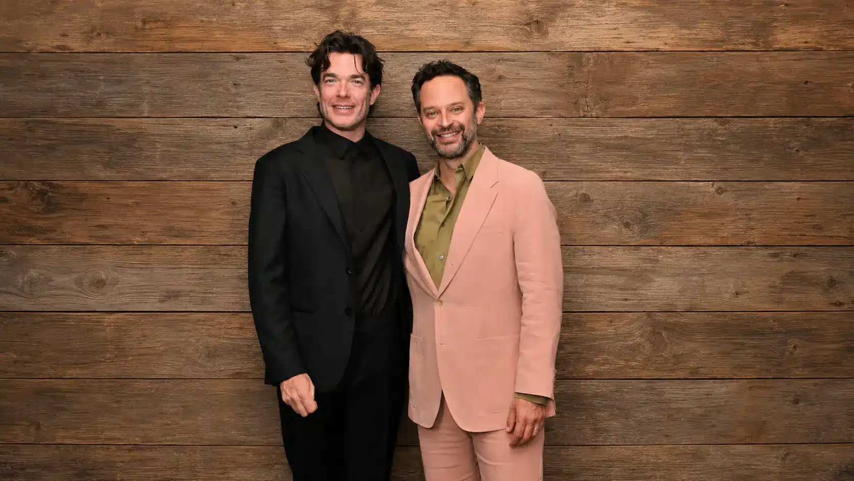 john mulaney and nick kroll men in formal suits standing together before a rustic wooden plank background