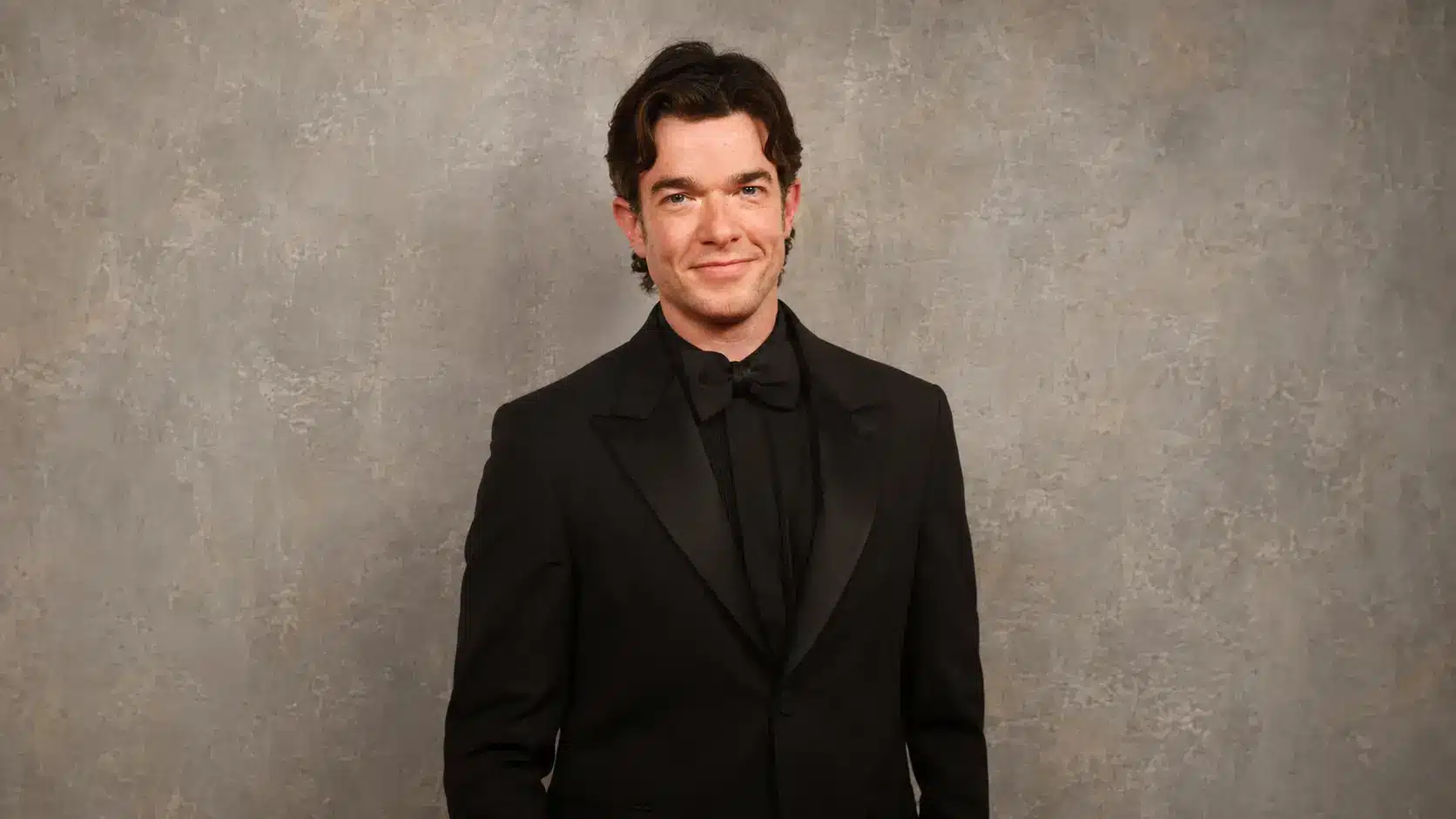 john mulaney in a black formal suit smiling against a gray rustic textured background in a clean portrait setting
