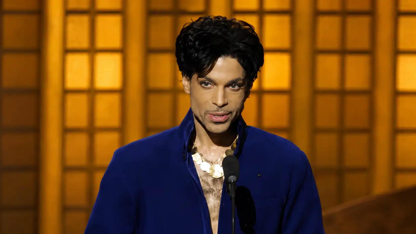 male musician in blue outfit standing at microphone before a golden paneled stage backdrop in a wide frame