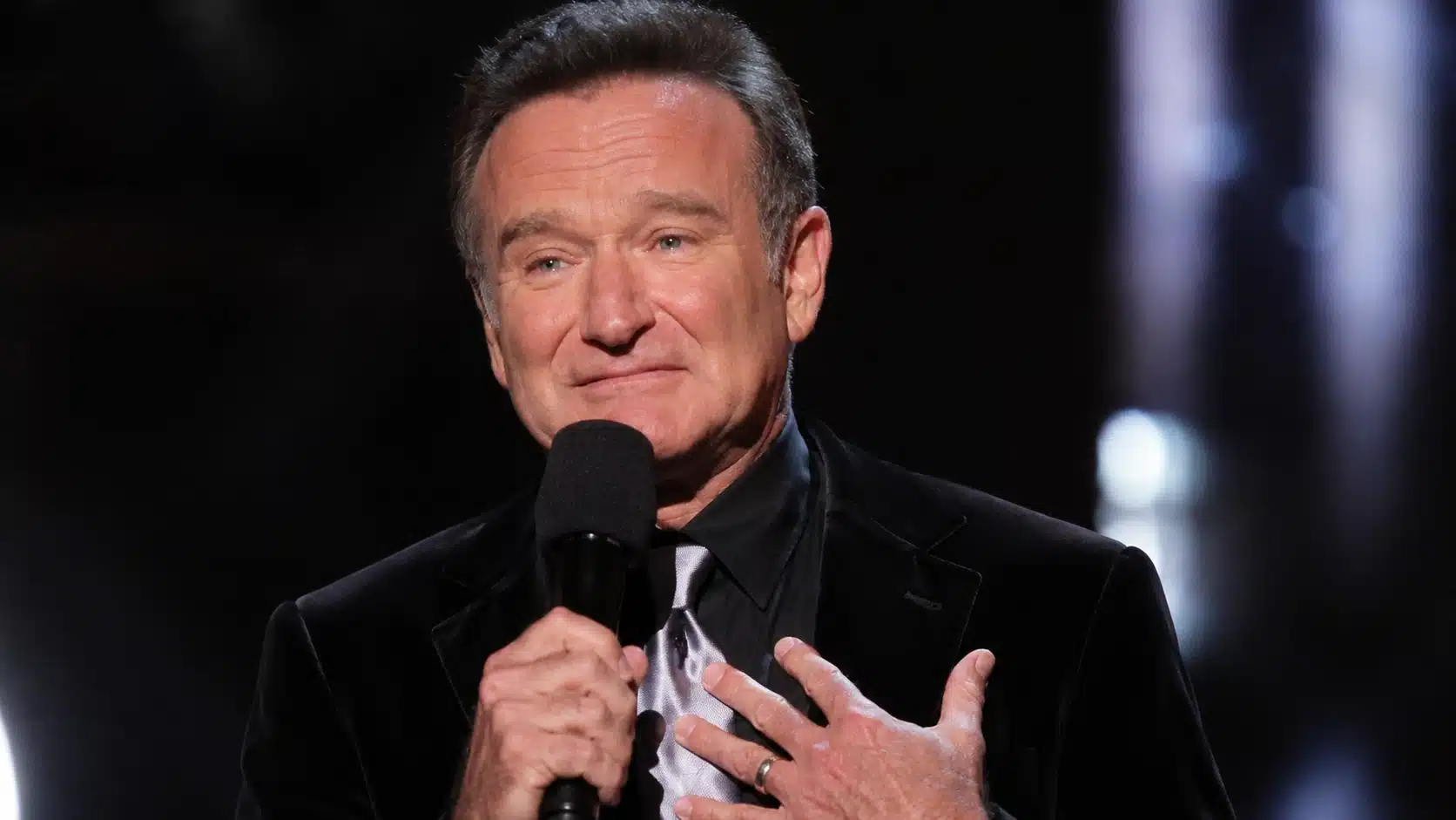 man delivering an emotional speech on stage while holding a microphone, wearing a dark suit under dramatic spotlight