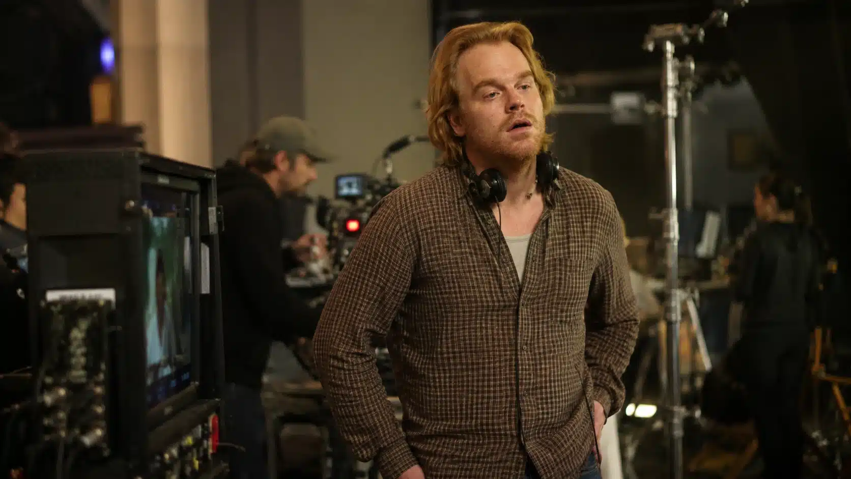 man in casual shirt standing in a busy filming room with headphones, cameras, monitors, crew members, and studio lights