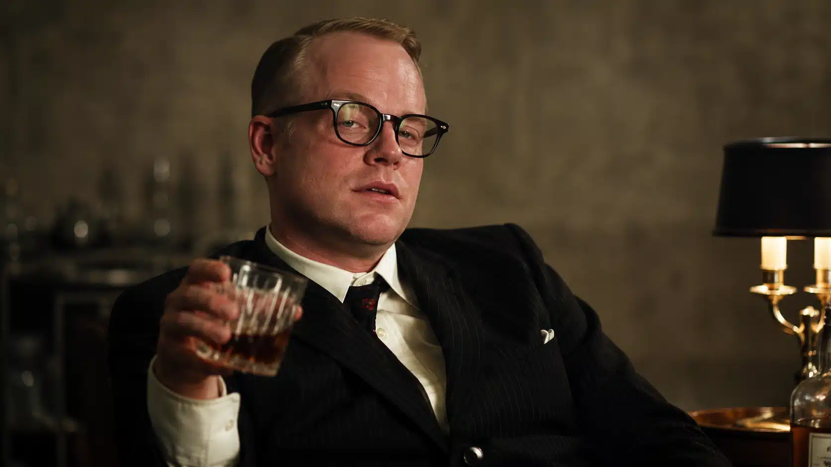 man in pinstripe suit holding a drink, looking slightly drunk in a dim room with warm lamp lighting