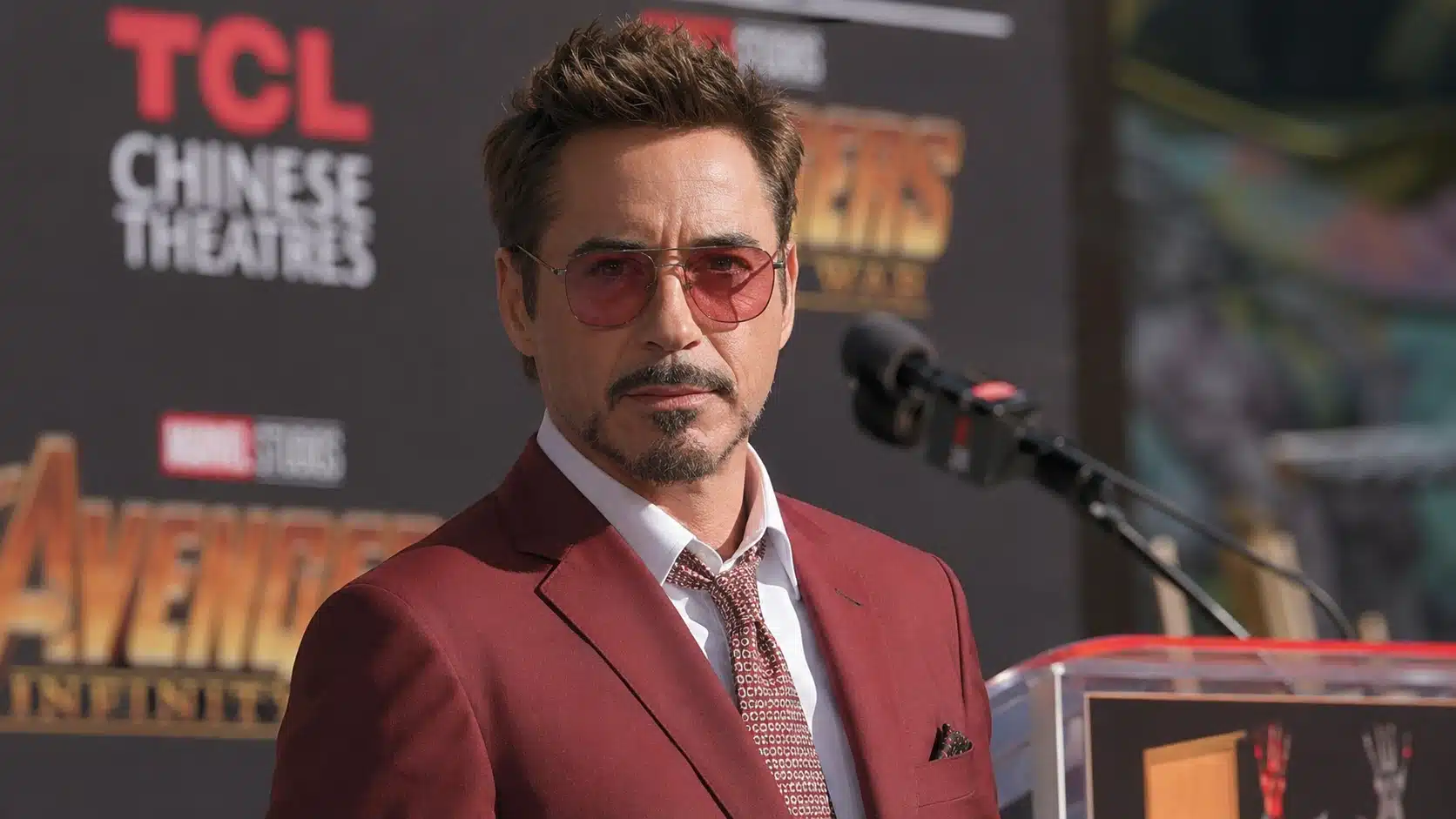 man in red suit standing at a sunny press event stage with microphones, looking at camera against a branded backdrop