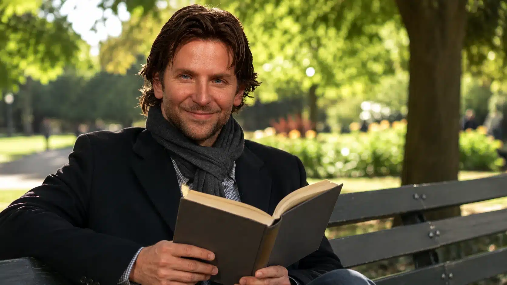 man smiling at camera while sitting on a park bench in daylight, holding an open book with green trees behind him
