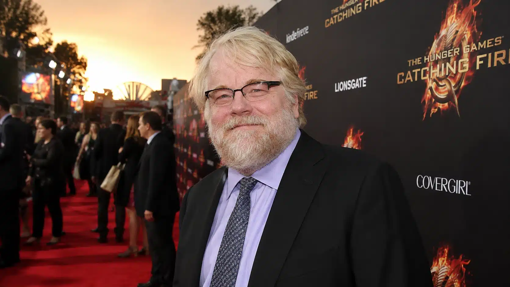 man smiling on a red carpet at sunset during a movie premiere, wearing a dark suit near event backdrop