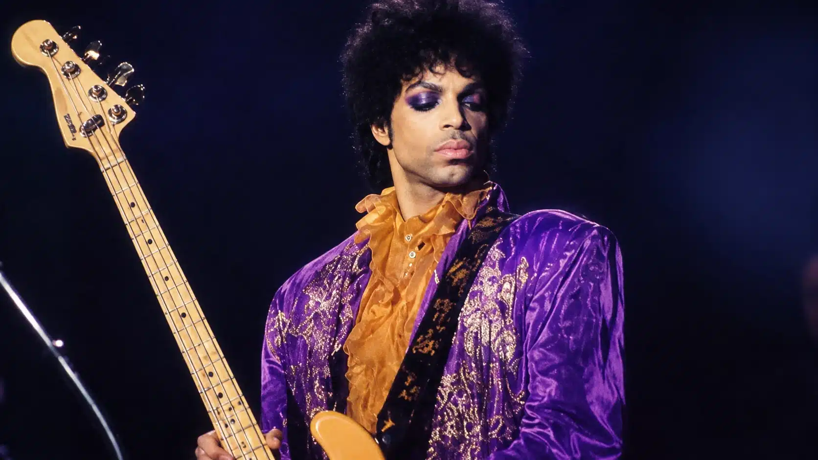 musician in purple jacket and orange ruffled shirt plays bass guitar on stage against a dark, concert background