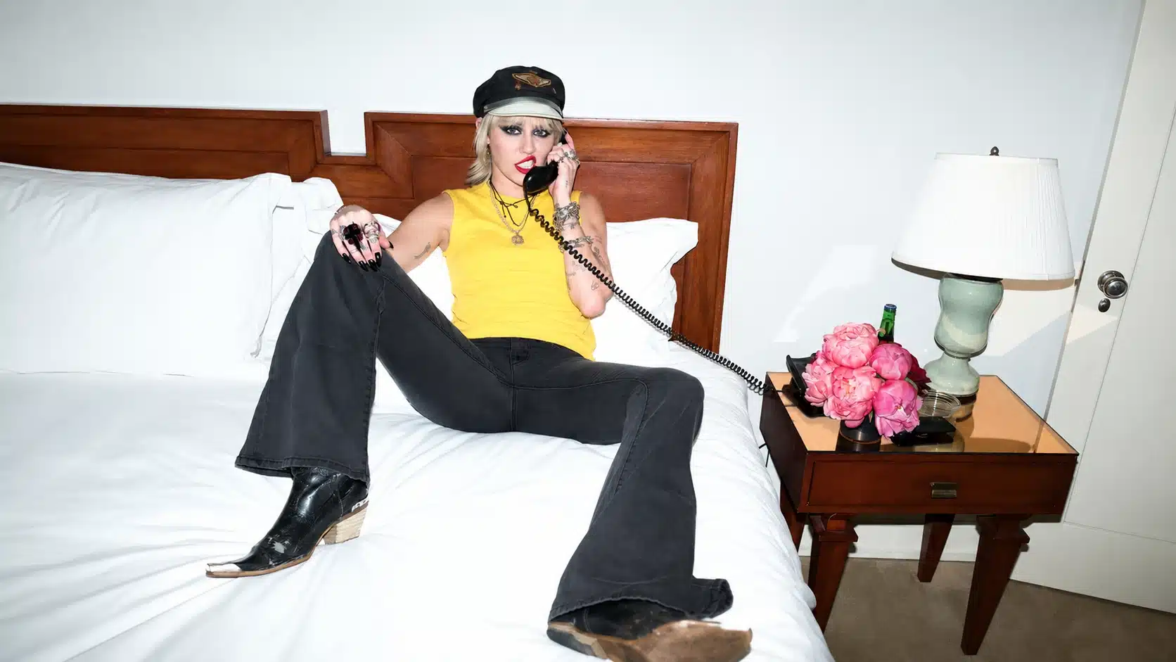 person lounging on white bed in yellow tank, black jeans, boots, and cap, holding vintage phone beside pink flowers