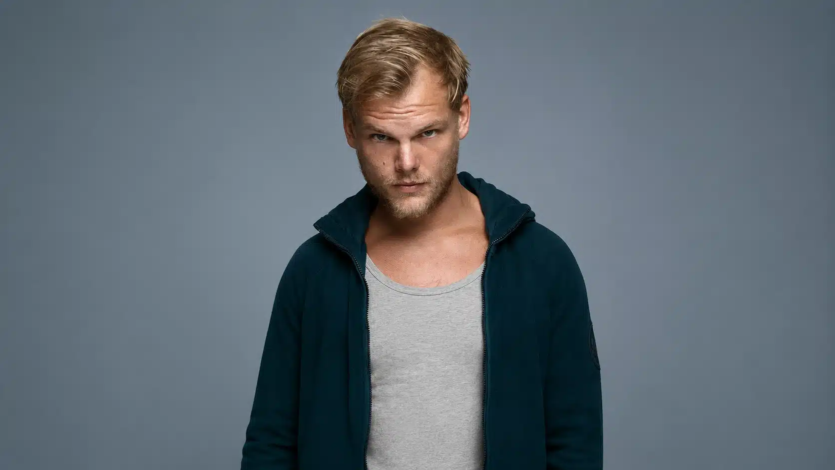 serious blond man in dark teal jacket and gray shirt against a muted blue-gray studio background, facing camera