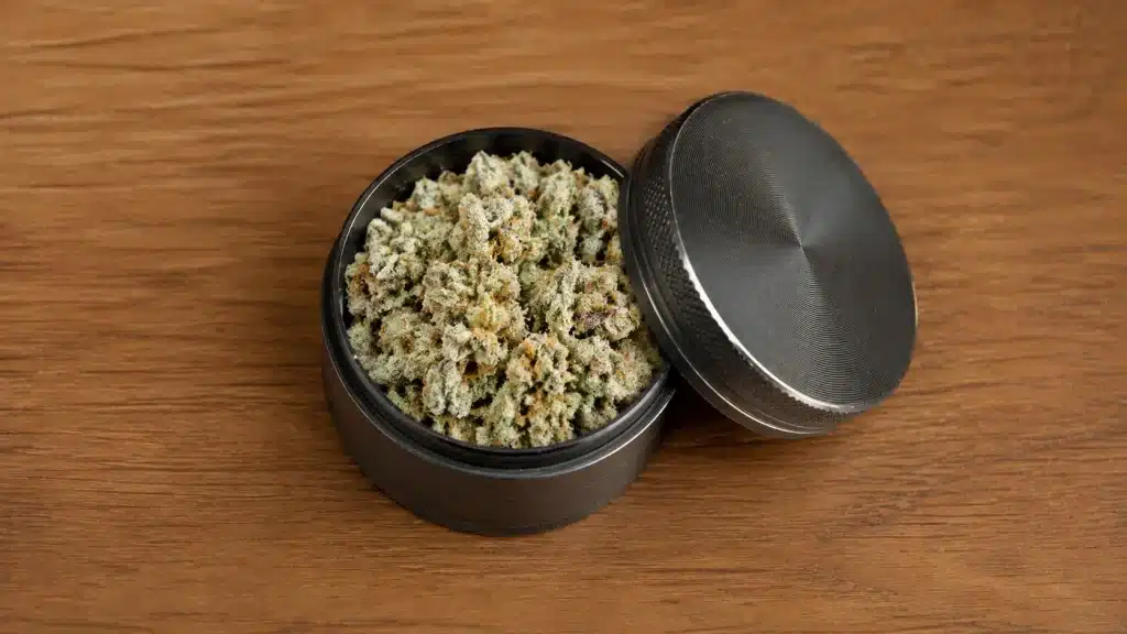 small crushed cannabis buds sit inside an open black metal grinder on a warm wooden table beside its textured lid