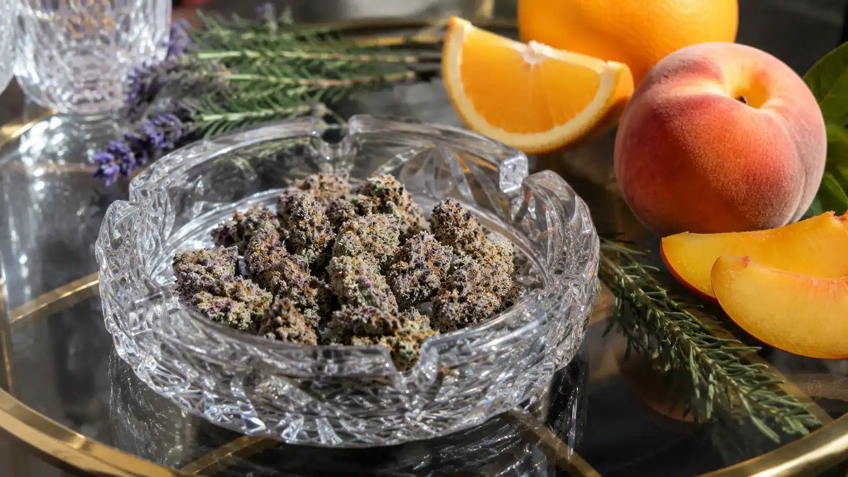 small violet cannabis buds in crystal ashtray with peach, orange, pine, and lavender on a reflective glass table
