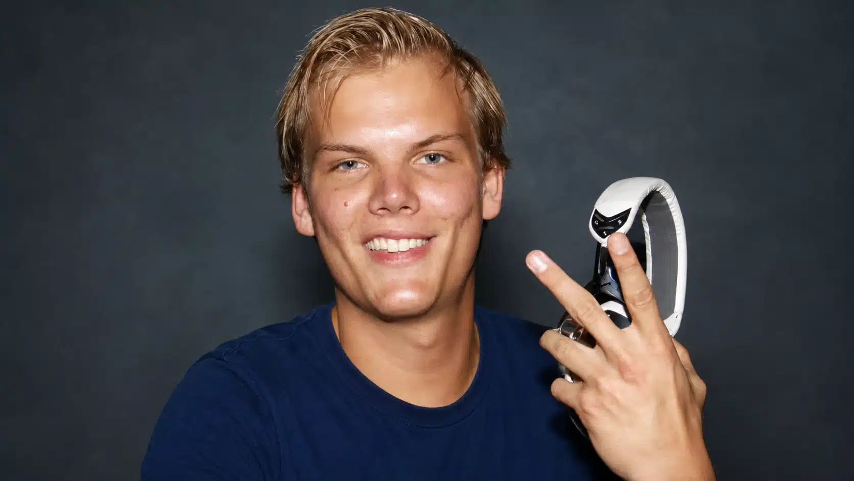 smiling blond man in navy shirt holding white headphones and making peace sign against dark charcoal studio background