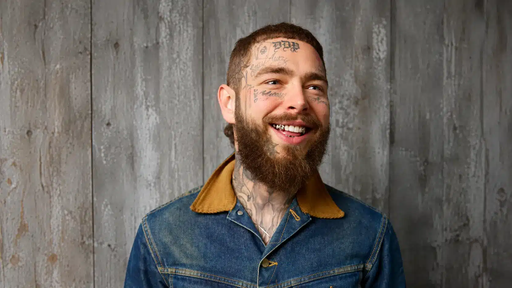 smiling post malone with face tattoos wearing a denim jacket against a gray rustic wooden wall background