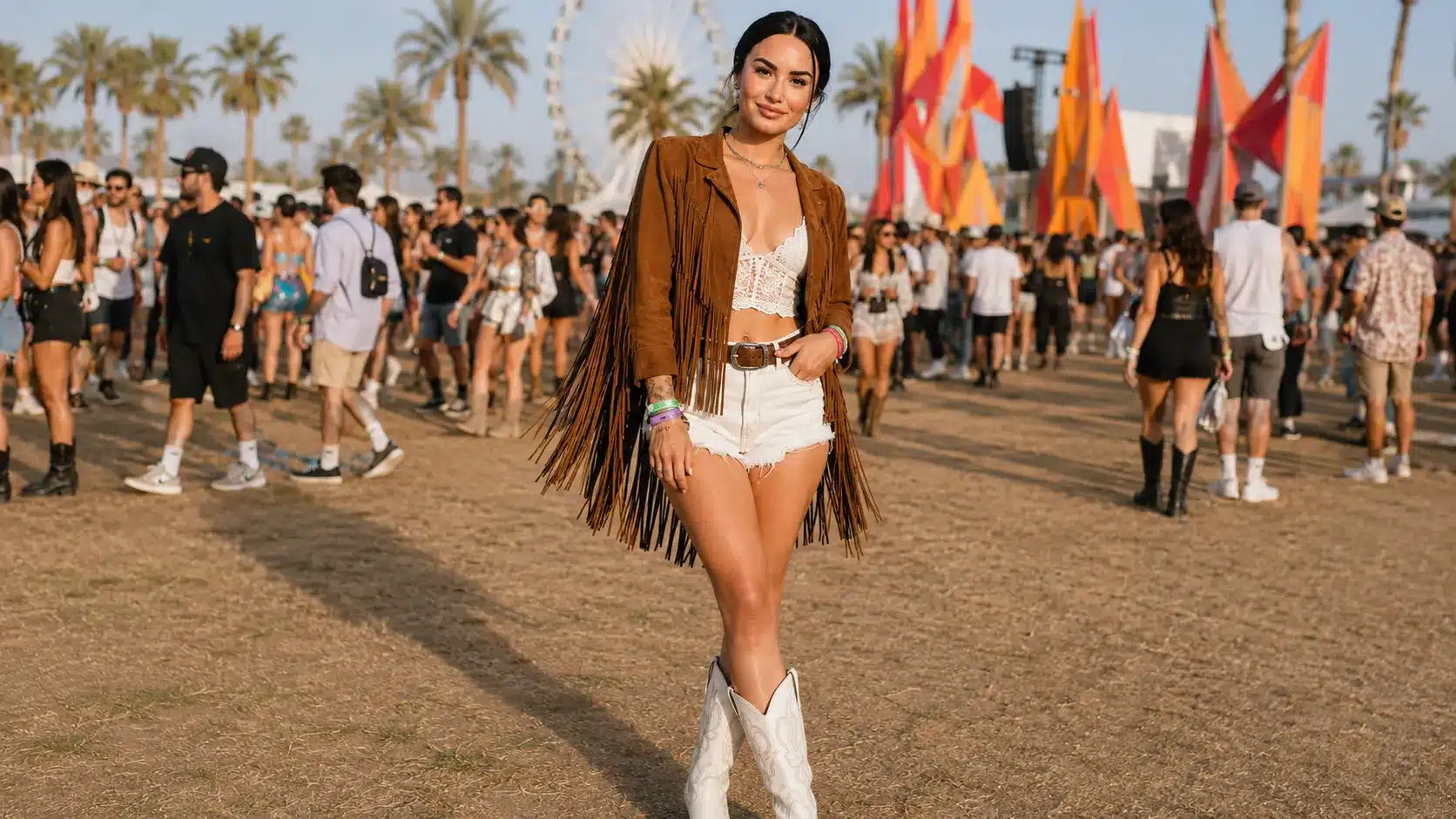 smiling woman in fringe jacket and cowboy boots poses at sunny music festival with crowd and palm trees nearby behind