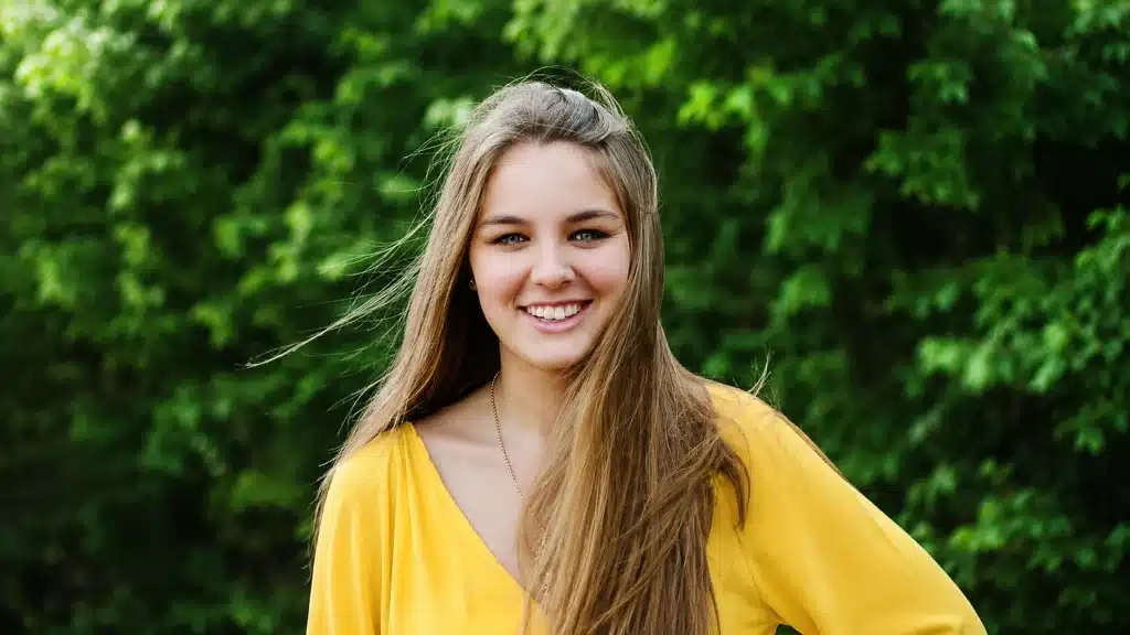 smiling young woman in bright yellow blouse stands outdoors before softly blurred green trees, with natural light