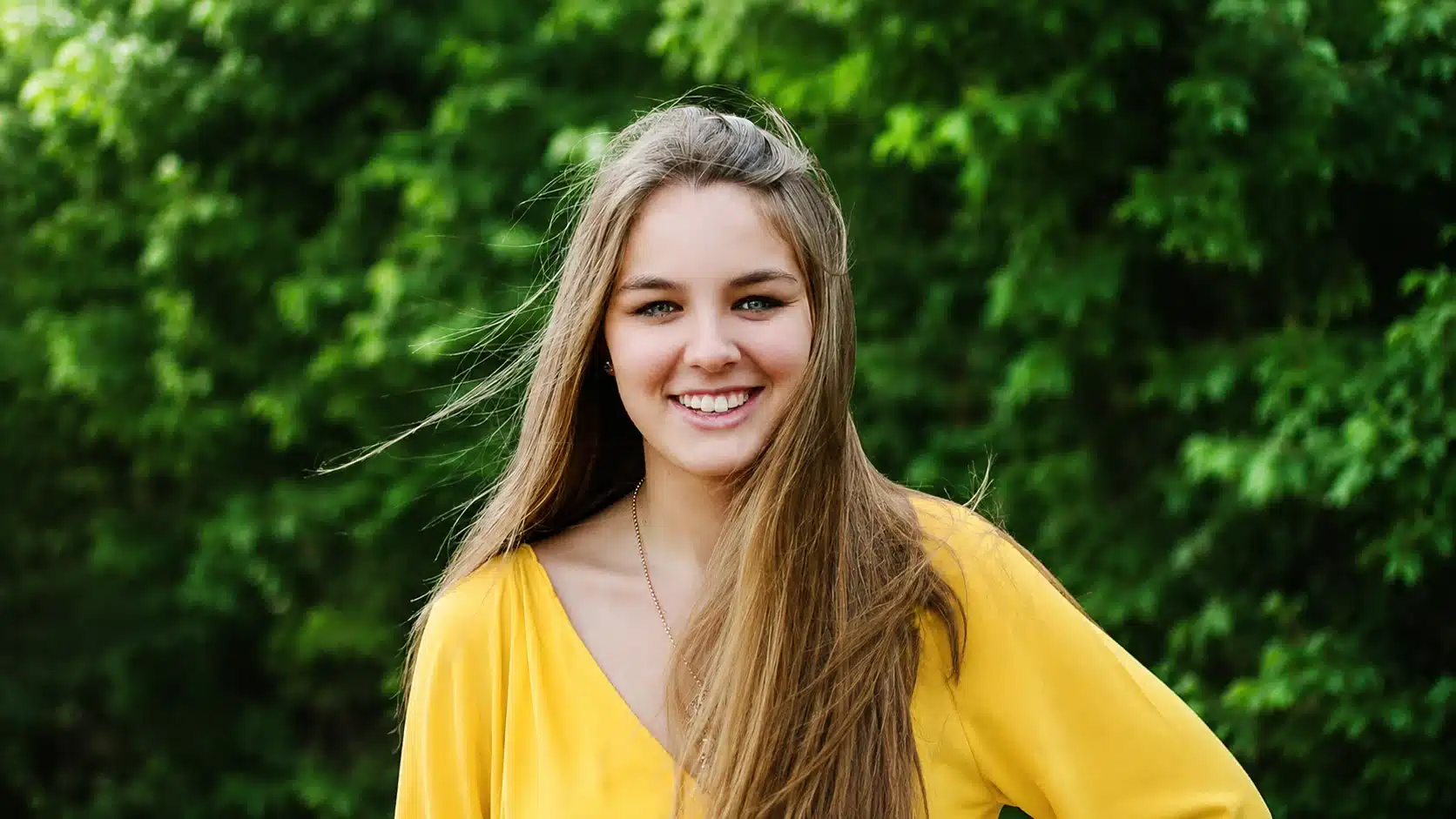 smiling young woman in bright yellow blouse stands outdoors before softly blurred green trees, with natural light