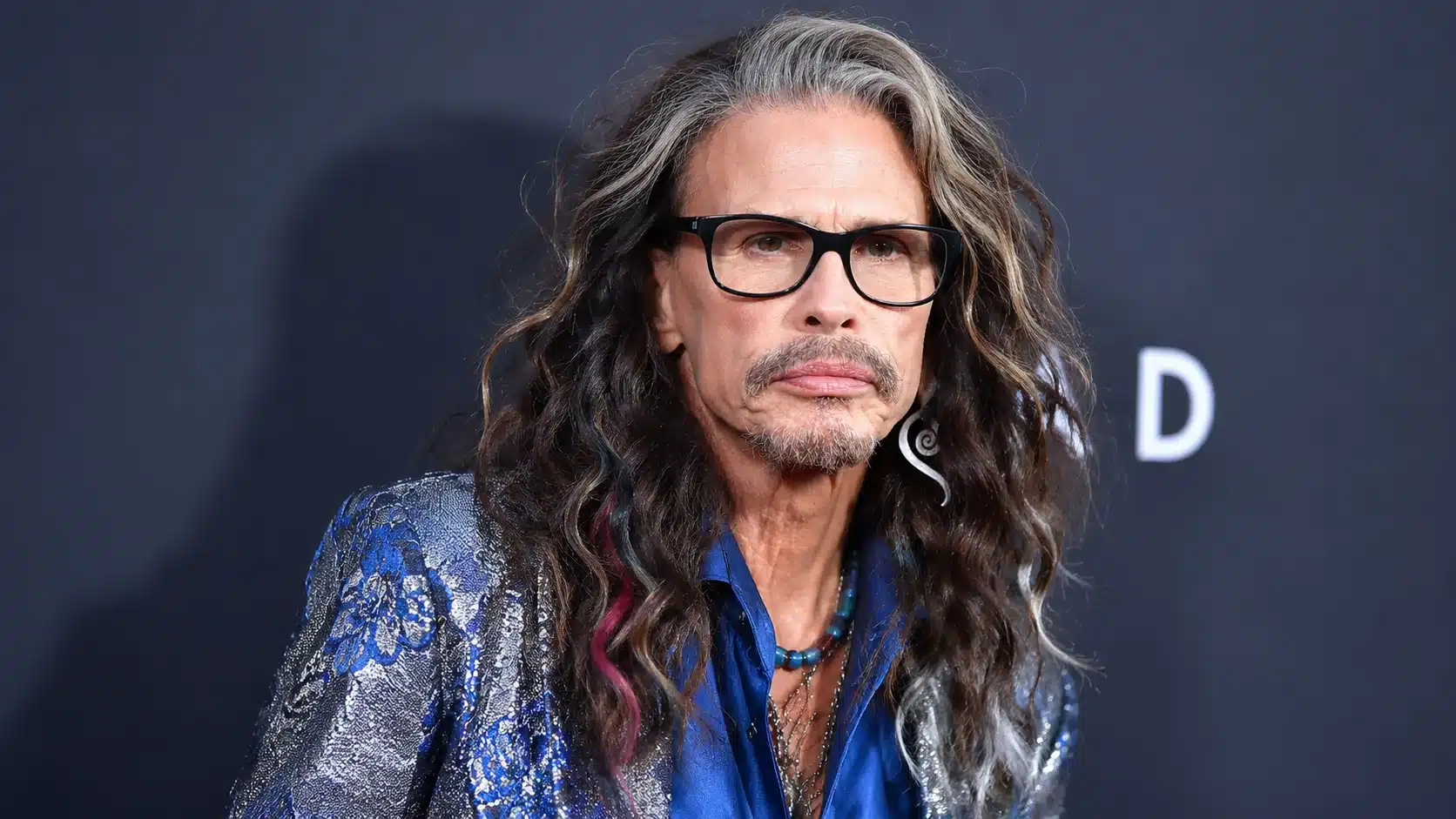 steven tyler portrait with open hair, glasses, blue shirt, and silver coat against a dark blurred background