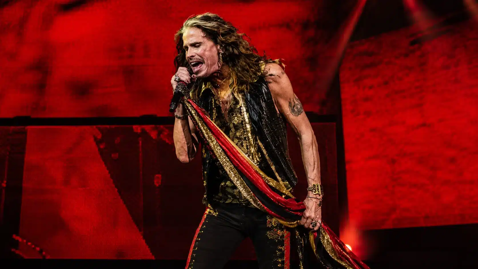 steven tyler singing on stage in black, gold, and red outfit, with bright red concert screen behind him