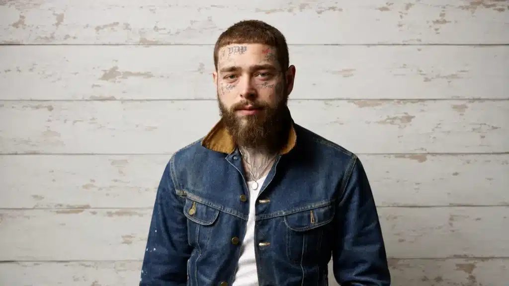 wide portrait of post malone with face tattoos and a denim jacket against a white rustic wooden wall background