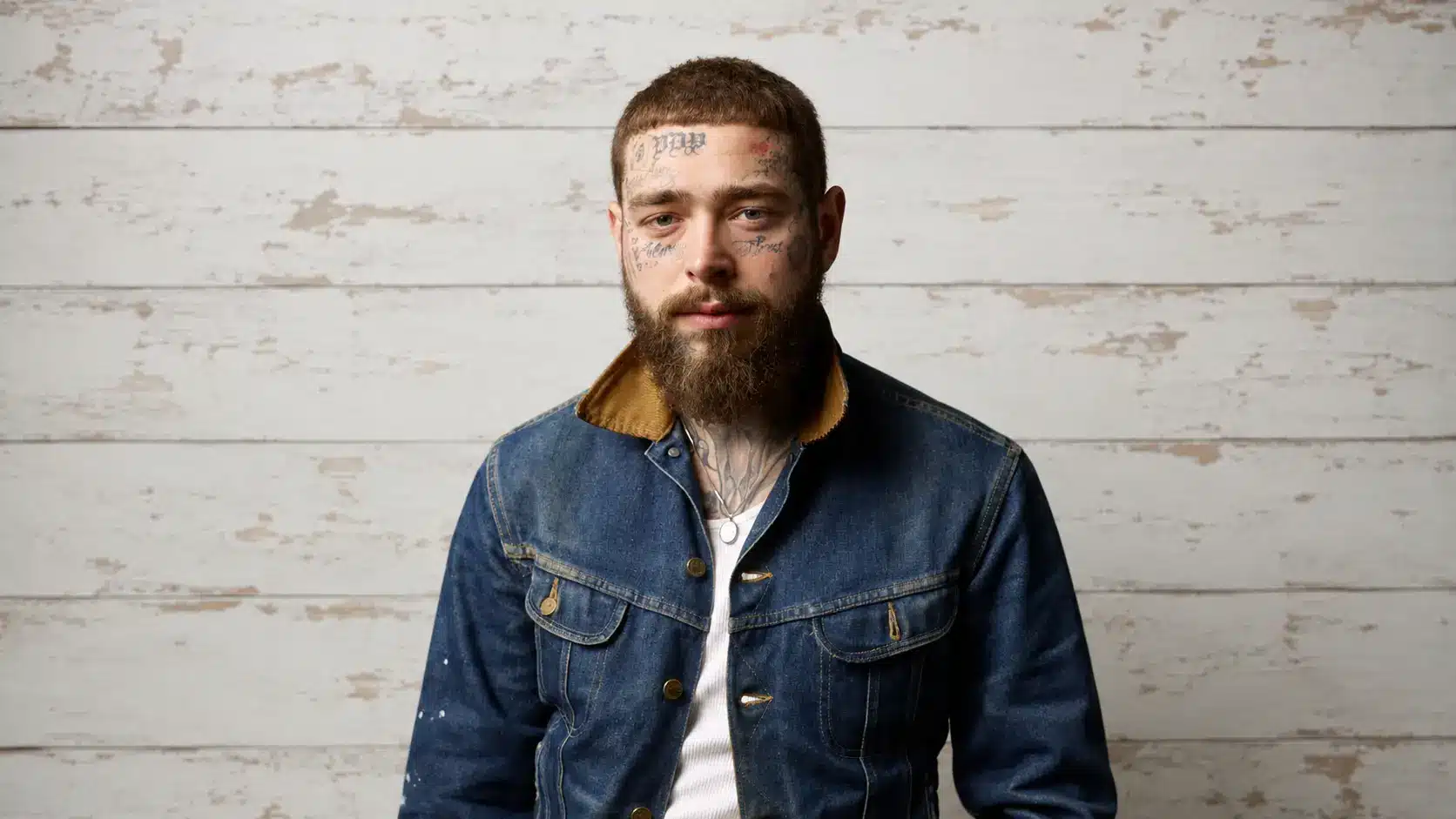wide portrait of post malone with face tattoos and a denim jacket against a white rustic wooden wall background