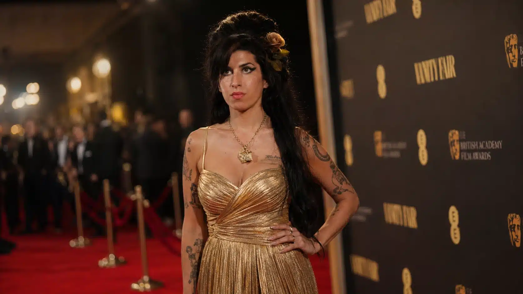 woman in a shimmering gold dress poses on a red carpet, wearing bold makeup and tattoos against a lit backdrop