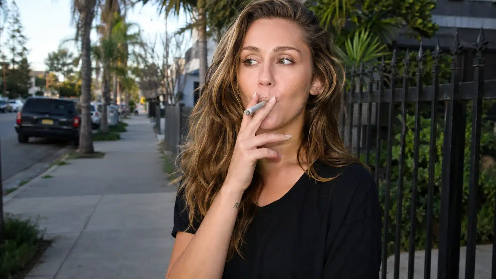 woman in black shirt smoking on a los angeles sidewalk with palm trees, parked cars, and metal fence behind her