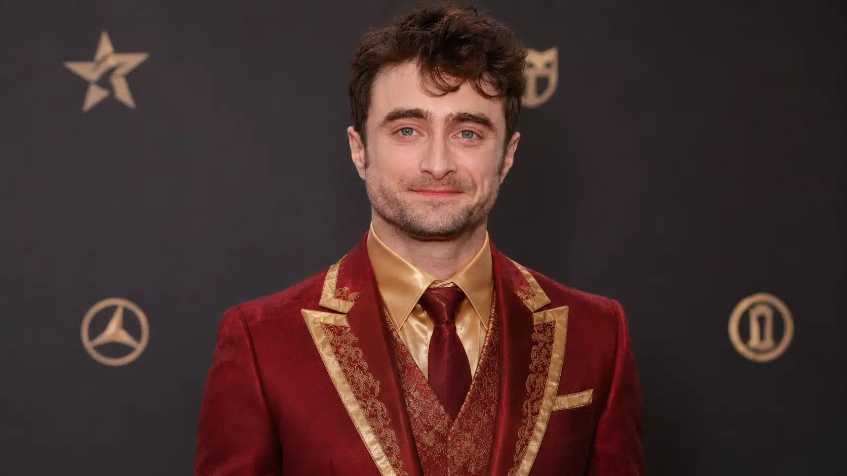 young man in a red and gold formal suit poses at a clean event backdrop with a few simple logos