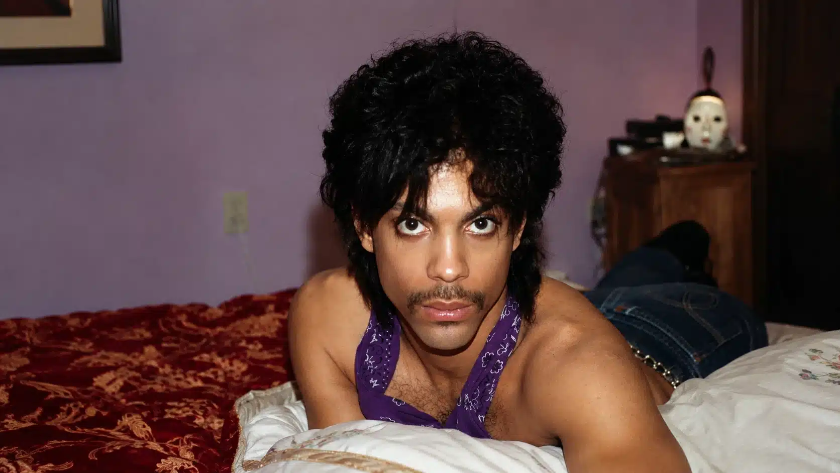 young man lying on bed in purple scarf, looking at camera, with lavender wall and red patterned bedding behind him