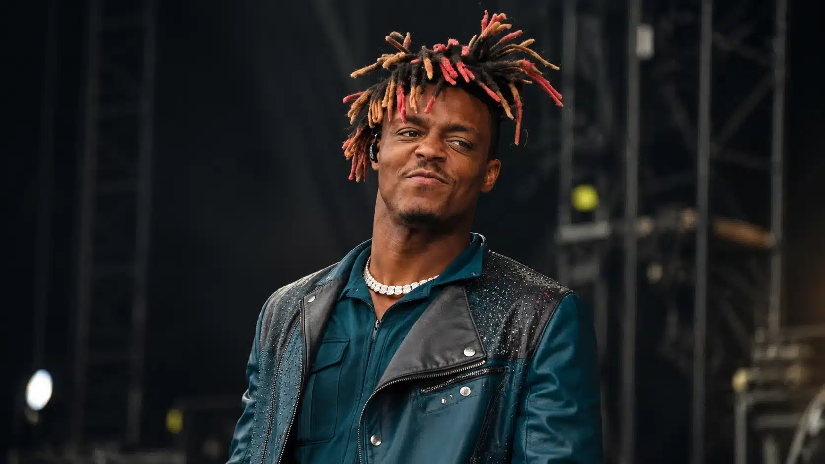 young performer in teal jacket and necklace smirking on a dark concert stage with colorful dreadlock and blurred rigging